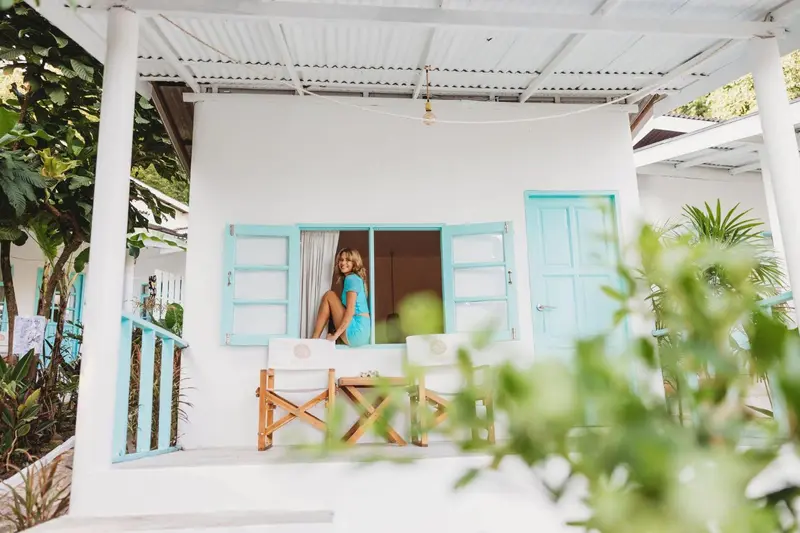 Une femme assise sur le rebord d'une fenêtre turquoise dans une maison blanche.