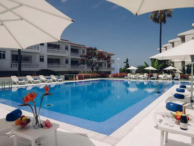 Une piscine bleue bordée de chaises longues et de parasols blancs, devant un bâtiment blanc.