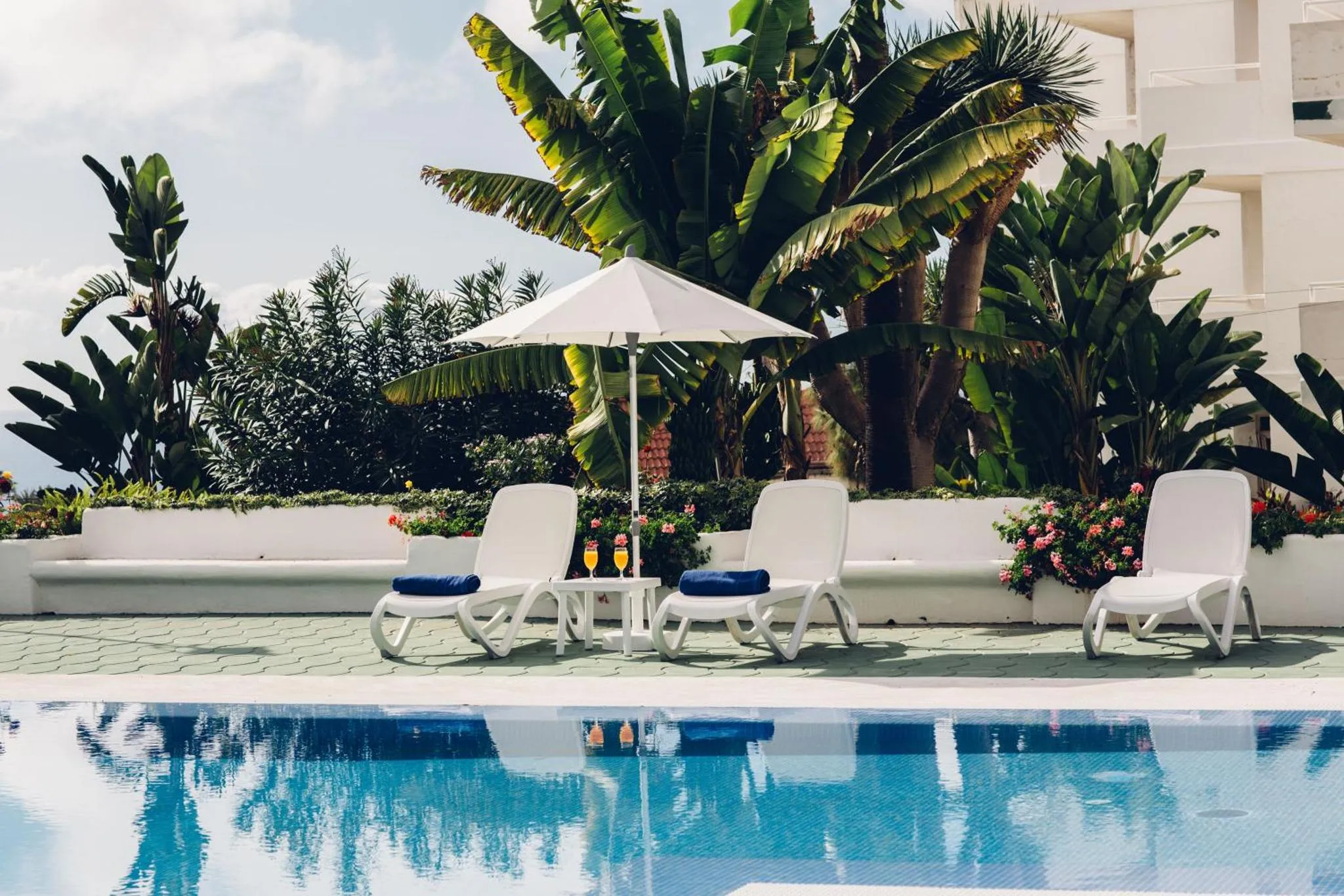 Transats blancs disposés au bord d'une piscine turquoise, ombragés par un parasol.