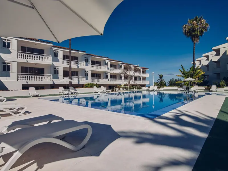 Une piscine rectangulaire bordée de transats blancs, devant un bâtiment blanc à balcons.