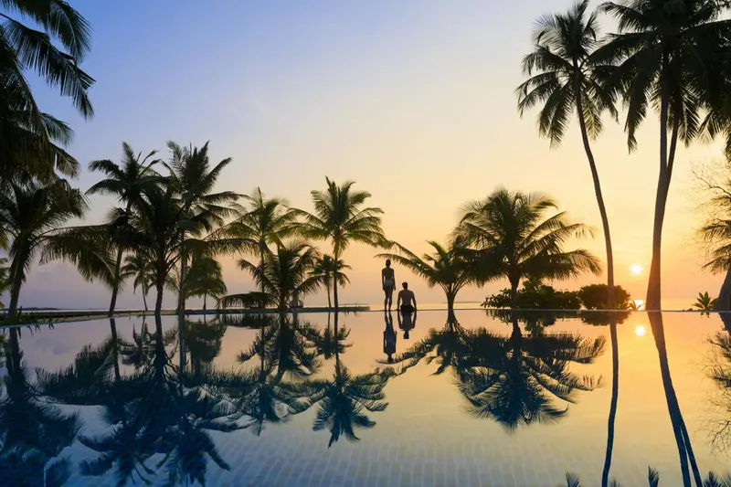 Une piscine à débordement reflète les palmiers et le ciel au coucher du soleil.