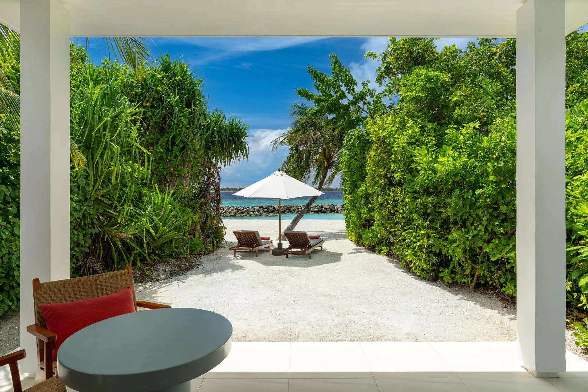 Vue sur une plage de sable blanc avec des transats et parasols depuis une terrasse ombragée.