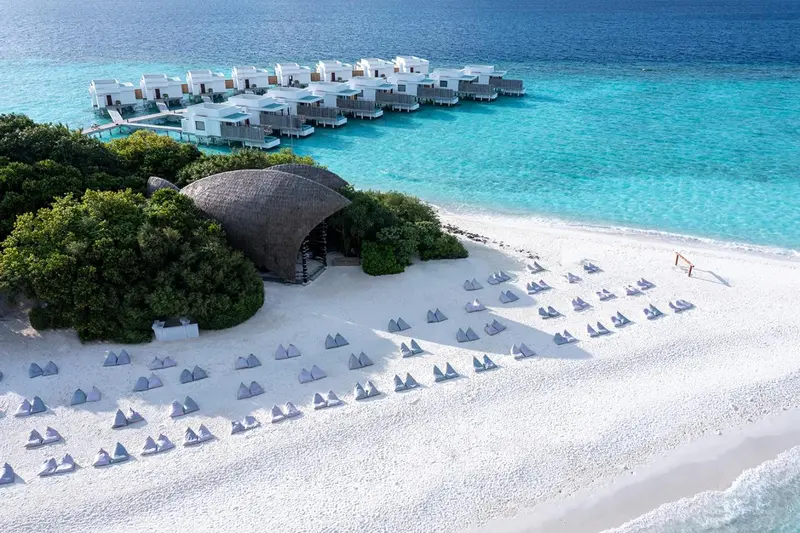 Vue aérienne d'une plage de sable blanc bordée de bungalows sur pilotis et de végétation luxuriante.