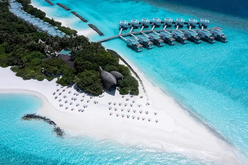 Vue aérienne d'une plage de sable blanc bordée de bungalows sur pilotis dans une eau turquoise.