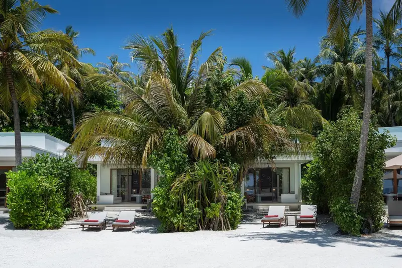 Des bungalows blancs bordent une plage de sable fin sous des palmiers.