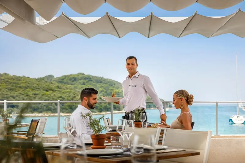 Un serveur présente un plat à deux clients assis à une table en terrasse avec vue sur la mer.