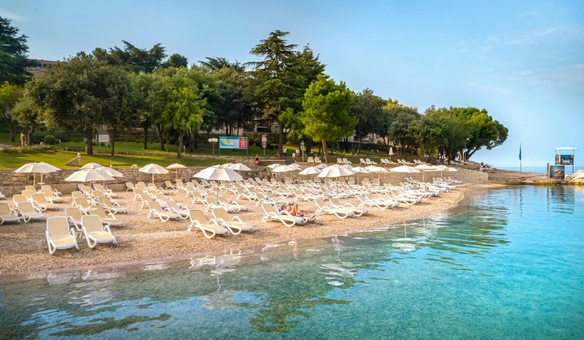 Transats blancs alignés sur une plage de galets au bord d'une eau turquoise.
