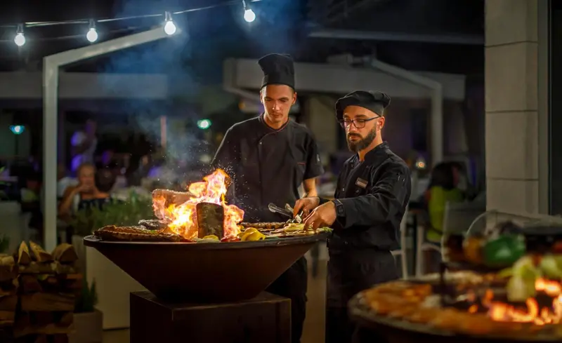 Deux cuisiniers préparent des grillades flambées devant un buffet extérieur.