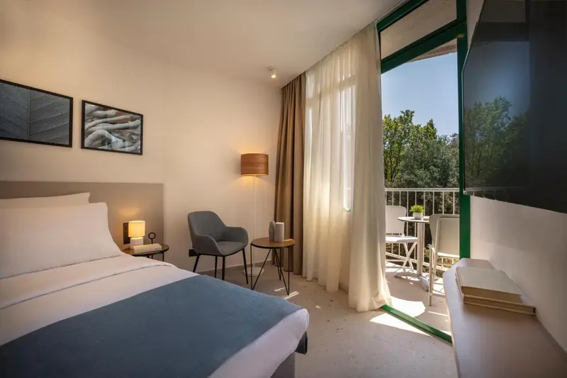Une chambre lumineuse avec un lit fait et un fauteuil près d'une baie vitrée ouverte sur une terrasse.