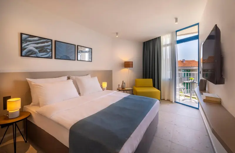 Chambre lumineuse avec un grand lit et un fauteuil jaune près d'une baie vitrée.