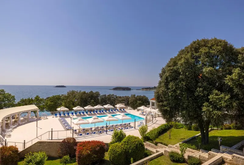Vue aérienne d'une piscine avec transats et parasols, bordée de végétation et surplombant la mer.