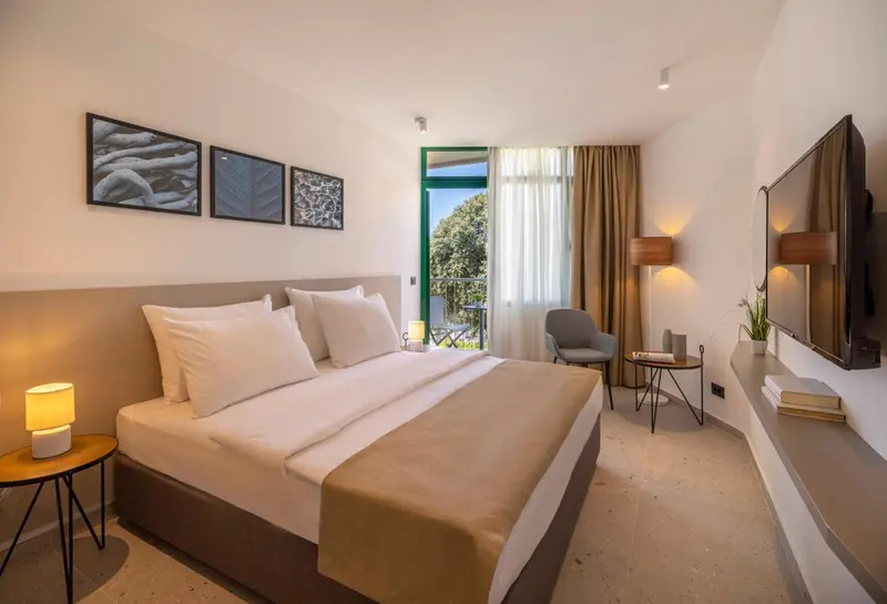 Chambre lumineuse avec un grand lit et un fauteuil près d'une porte-fenêtre.