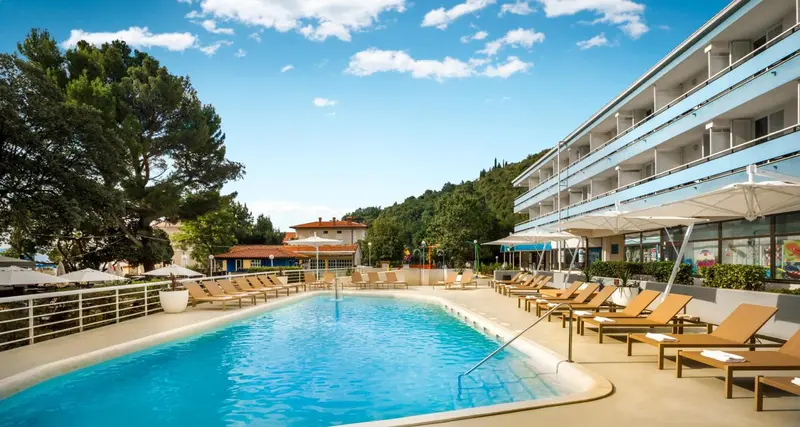 Une piscine bordée de transats devant un bâtiment blanc à plusieurs étages.