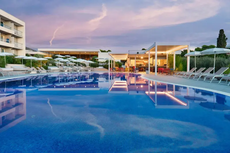 Piscine rectangulaire avec transats et un bar en arrière-plan au coucher du soleil.