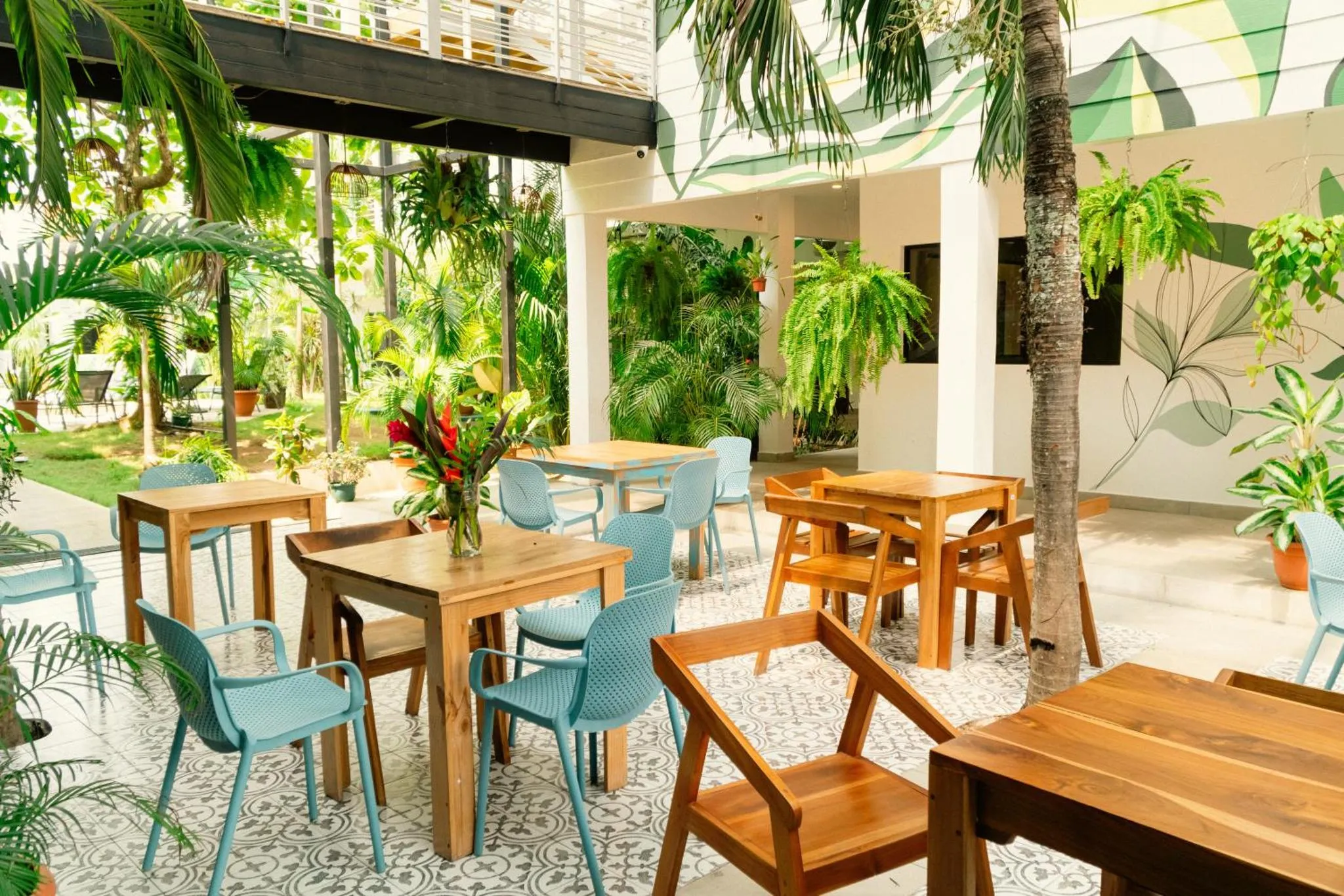 Tables et chaises en bois disposées sur une terrasse carrelée ombragée par des palmiers.