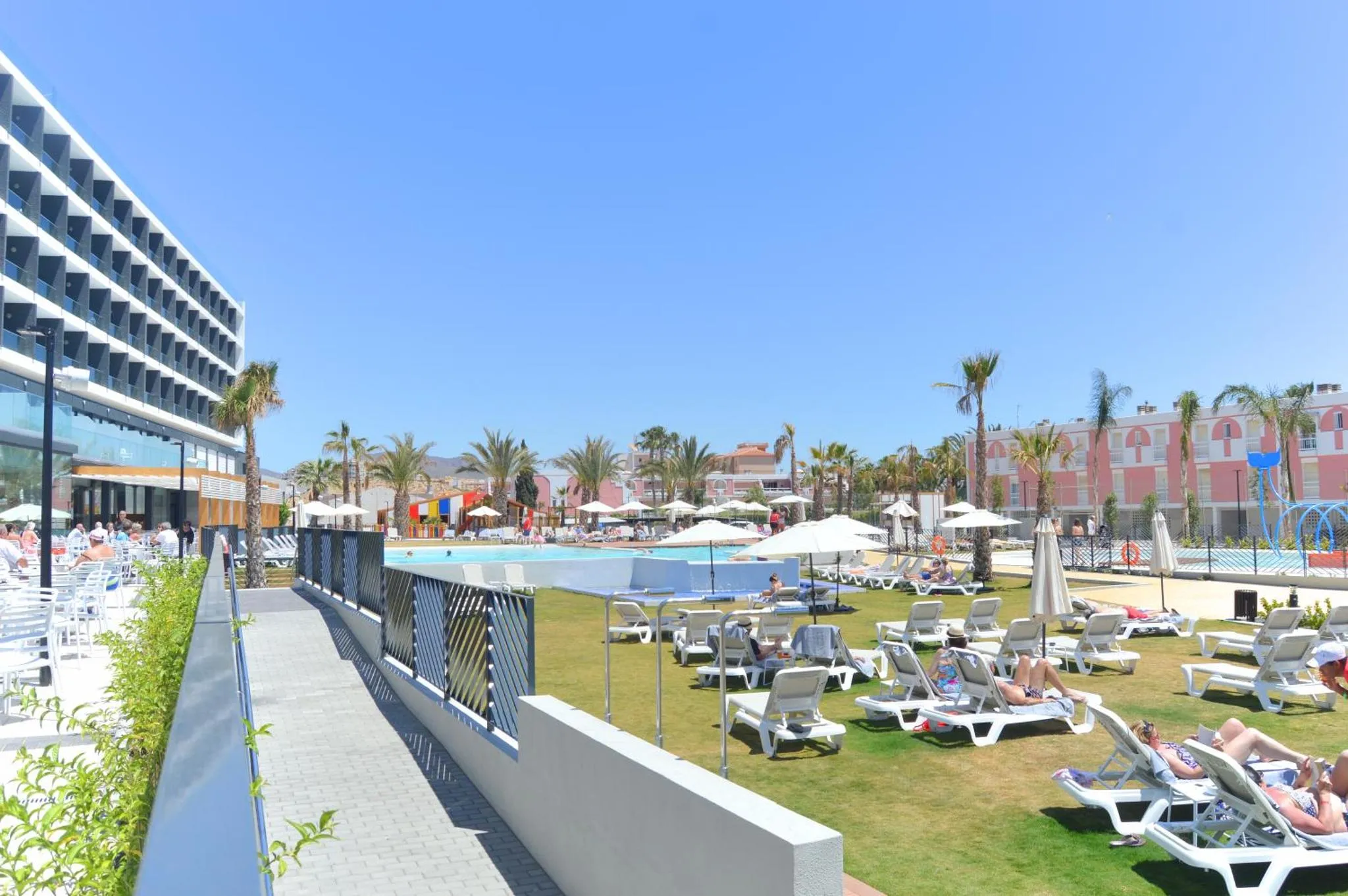 Transats alignés au bord d'une piscine ensoleillée, avec un bâtiment moderne en arrière-plan.