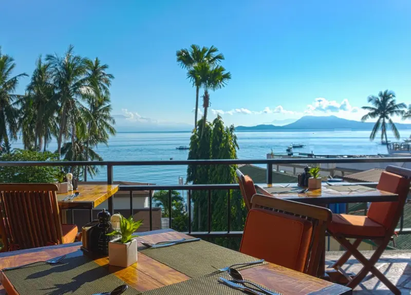 Tables de restaurant en extérieur avec vue sur la mer et les palmiers.