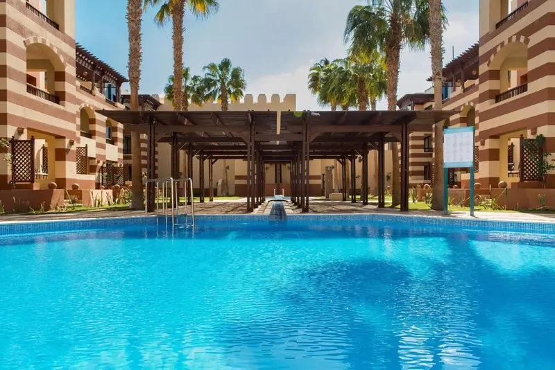 Une piscine turquoise bordée d'une terrasse ombragée et de bâtiments beiges.