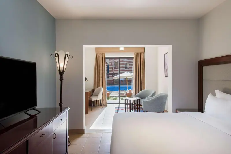 Une chambre lumineuse avec un lit et un espace salon donnant sur une piscine.