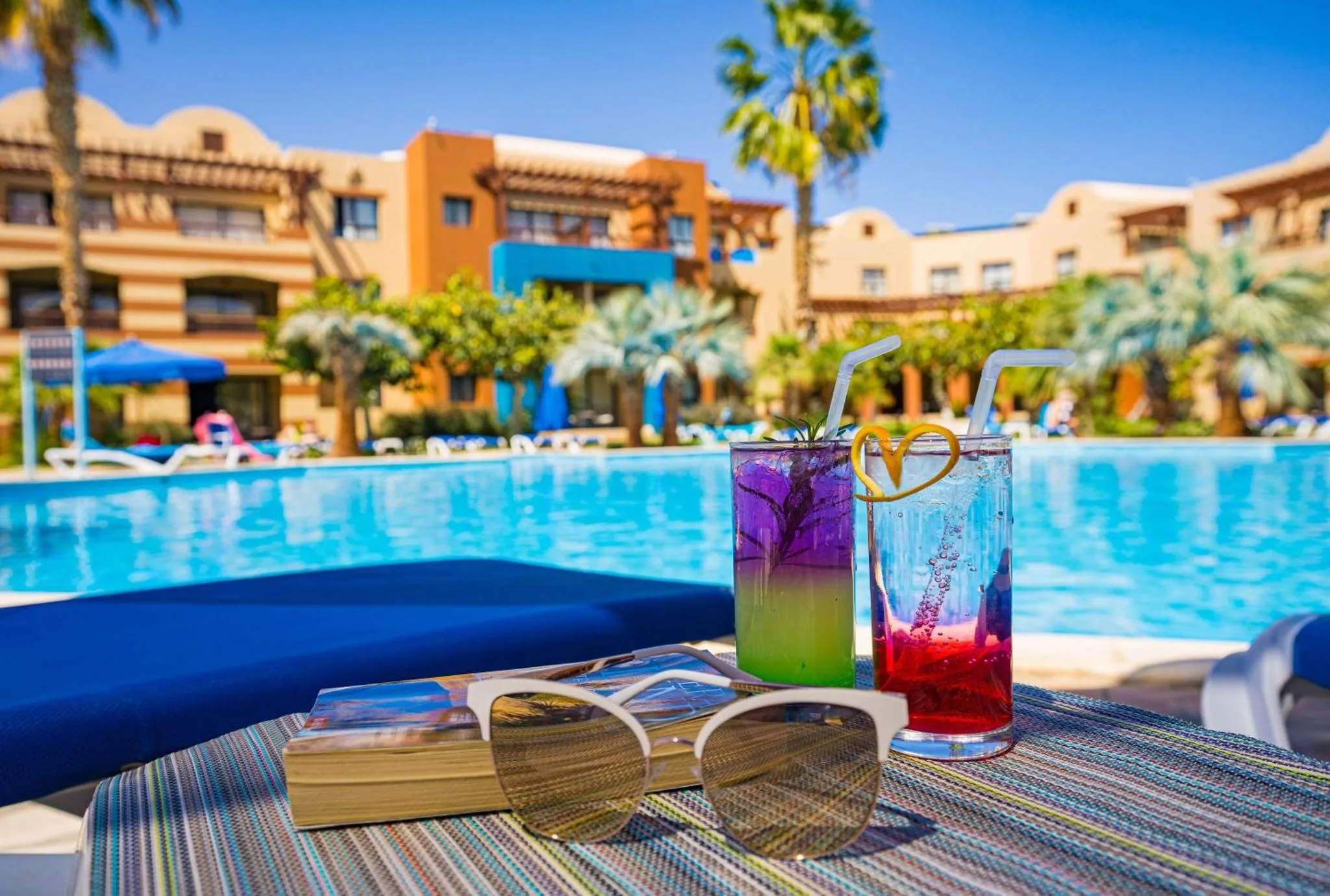 Deux boissons colorées reposent sur une table basse près d'une piscine ensoleillée.