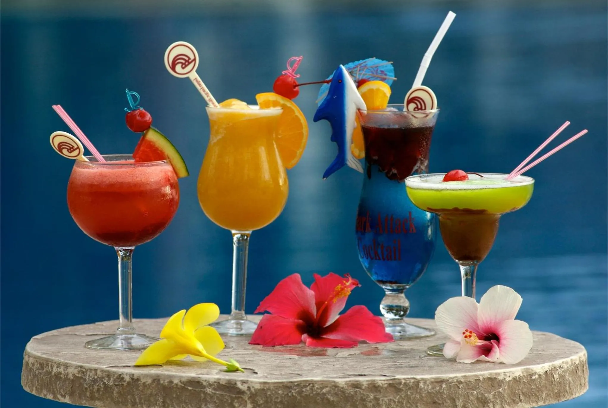 Des cocktails colorés et décorés de fruits sont présentés sur une table en pierre près d'une piscine.