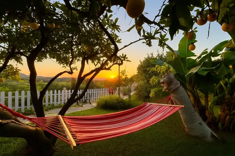 Un hamac rouge est suspendu entre deux arbres dans un jardin au coucher du soleil.