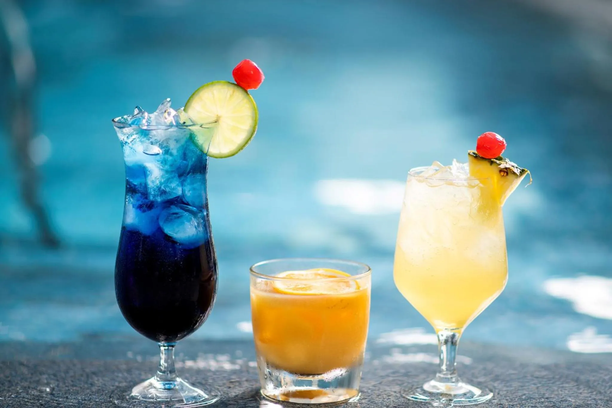 Trois cocktails colorés sont présentés sur une surface sombre, avec une piscine floue en arrière-plan.