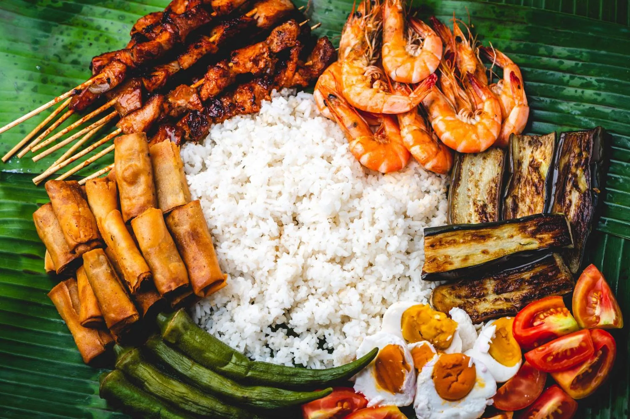 Assortiment de brochettes de viande, de fruits de mer, de riz et de légumes frais sur un lit de feuilles de bananier.