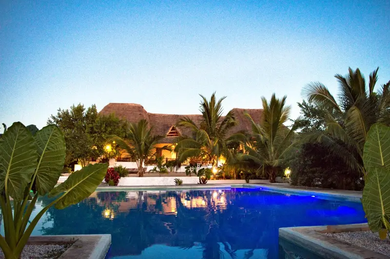 Une piscine illuminée reflète les lumières d'un bâtiment tropical au crépuscule.