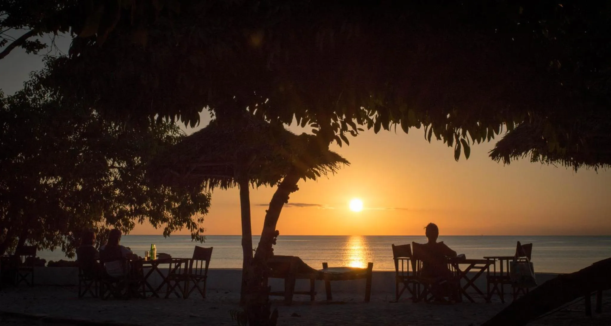 Des silhouettes sont assises à des tables au coucher du soleil sur une plage.