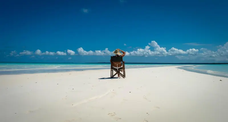 Une personne assise sur une chaise en bois face à une plage de sable blanc et une mer turquoise.