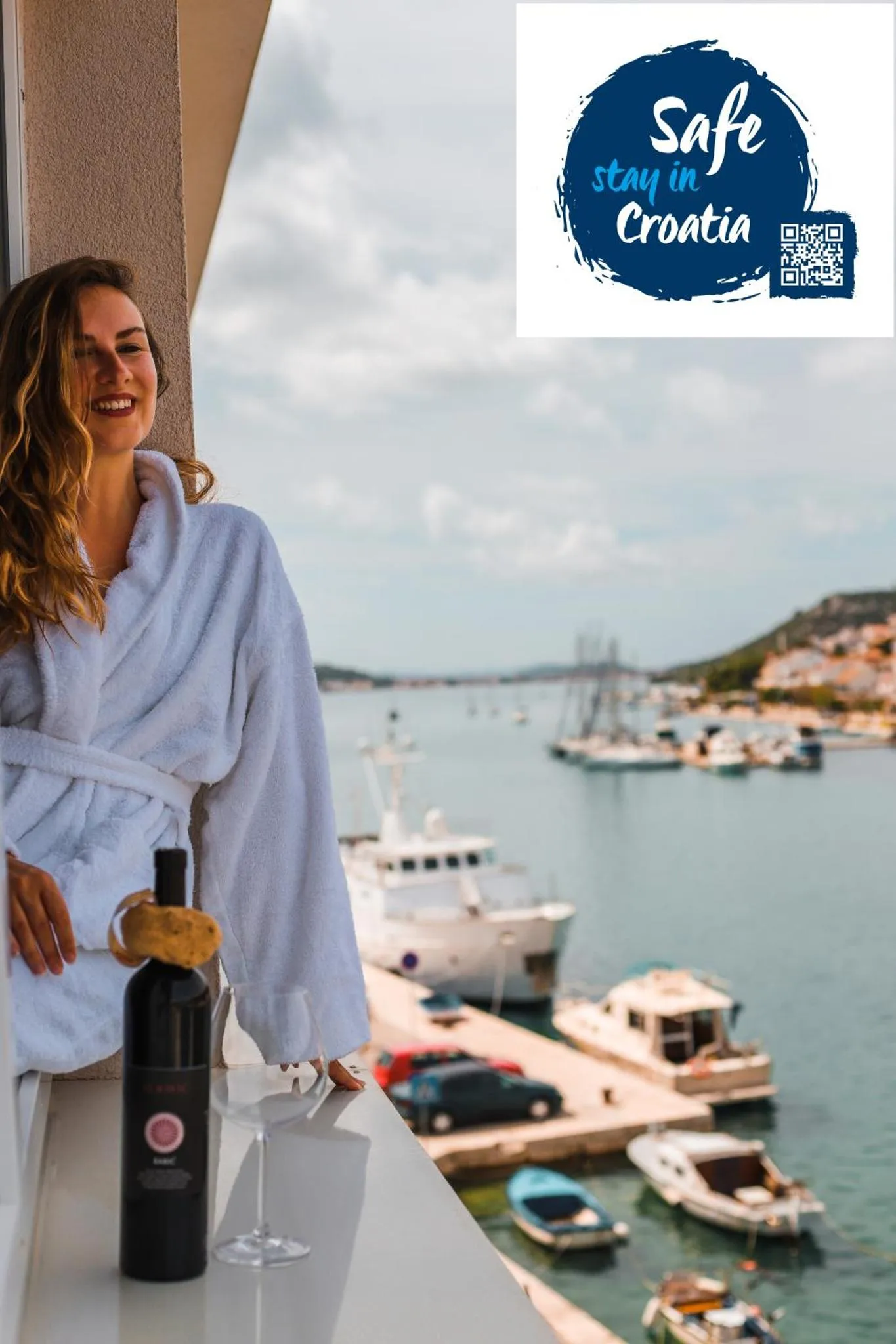 Une femme souriante en peignoir se tient sur une terrasse avec une bouteille de vin et un verre, surplombant un port de plaisance.