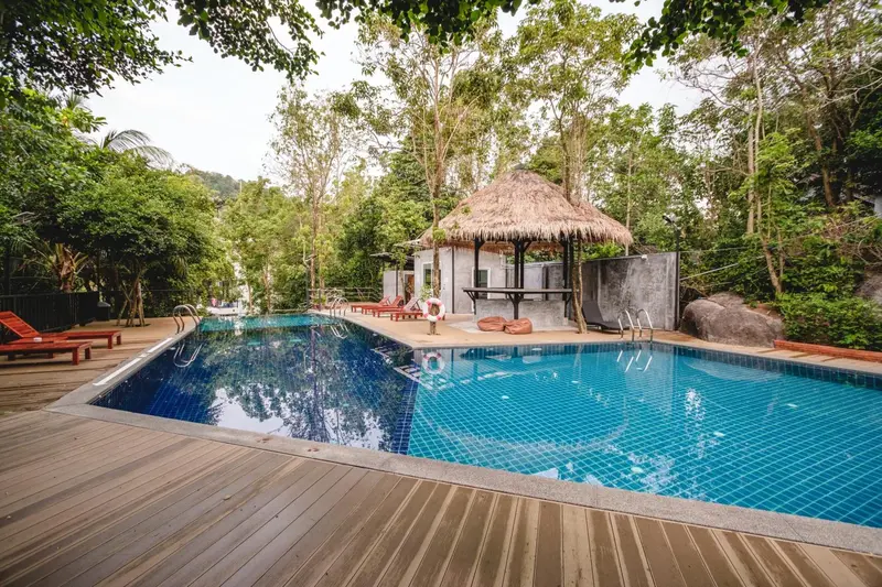 Une piscine rectangulaire bordée d'une terrasse en bois, avec un pavillon de style tropical en arrière-plan.