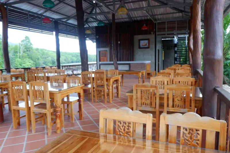 Une salle à manger en bois avec de nombreuses tables et chaises est ouverte sur une vue verdoyante.