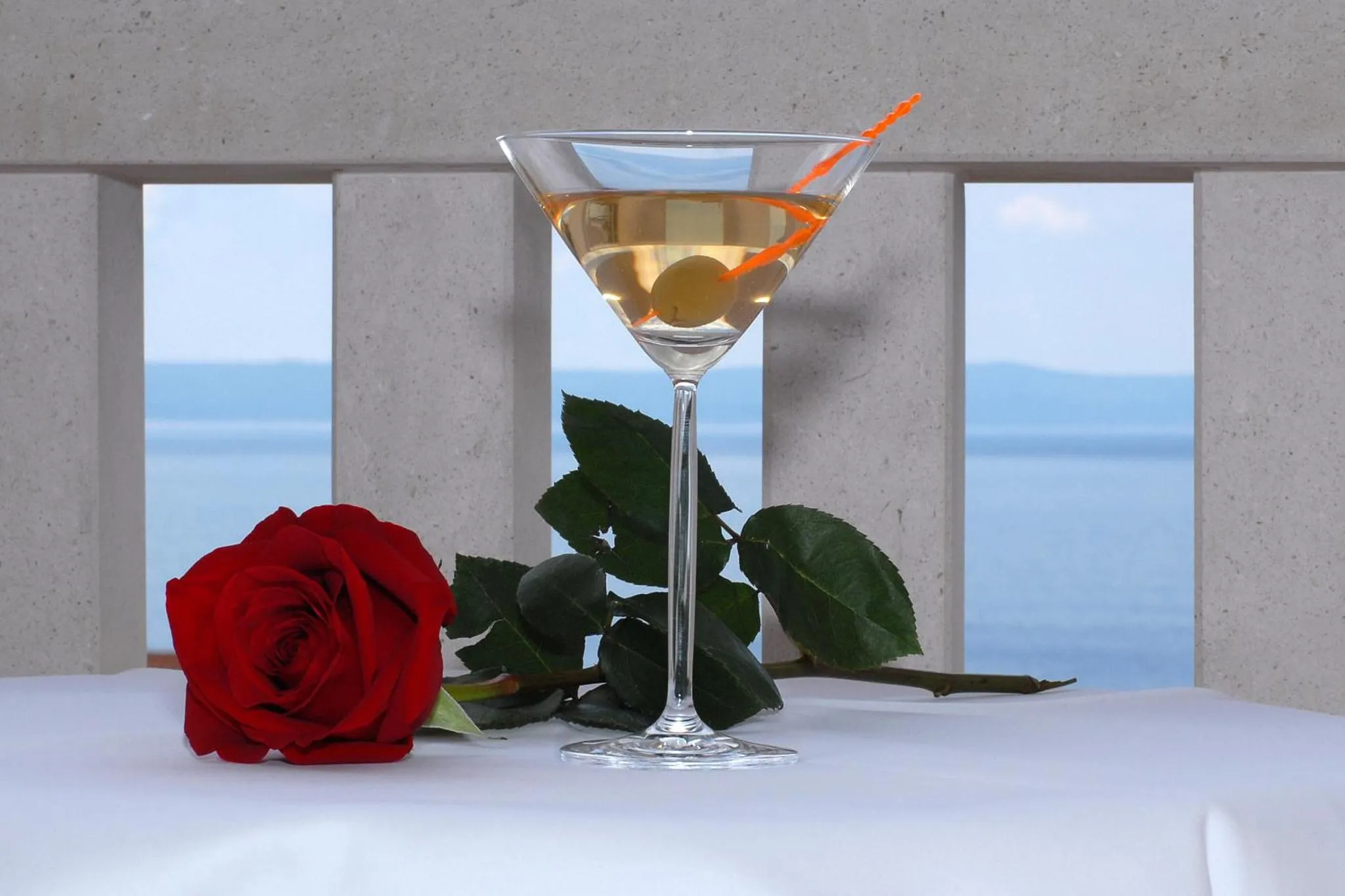 Un cocktail et une rose rouge sont posés sur une table blanche avec vue sur la mer.