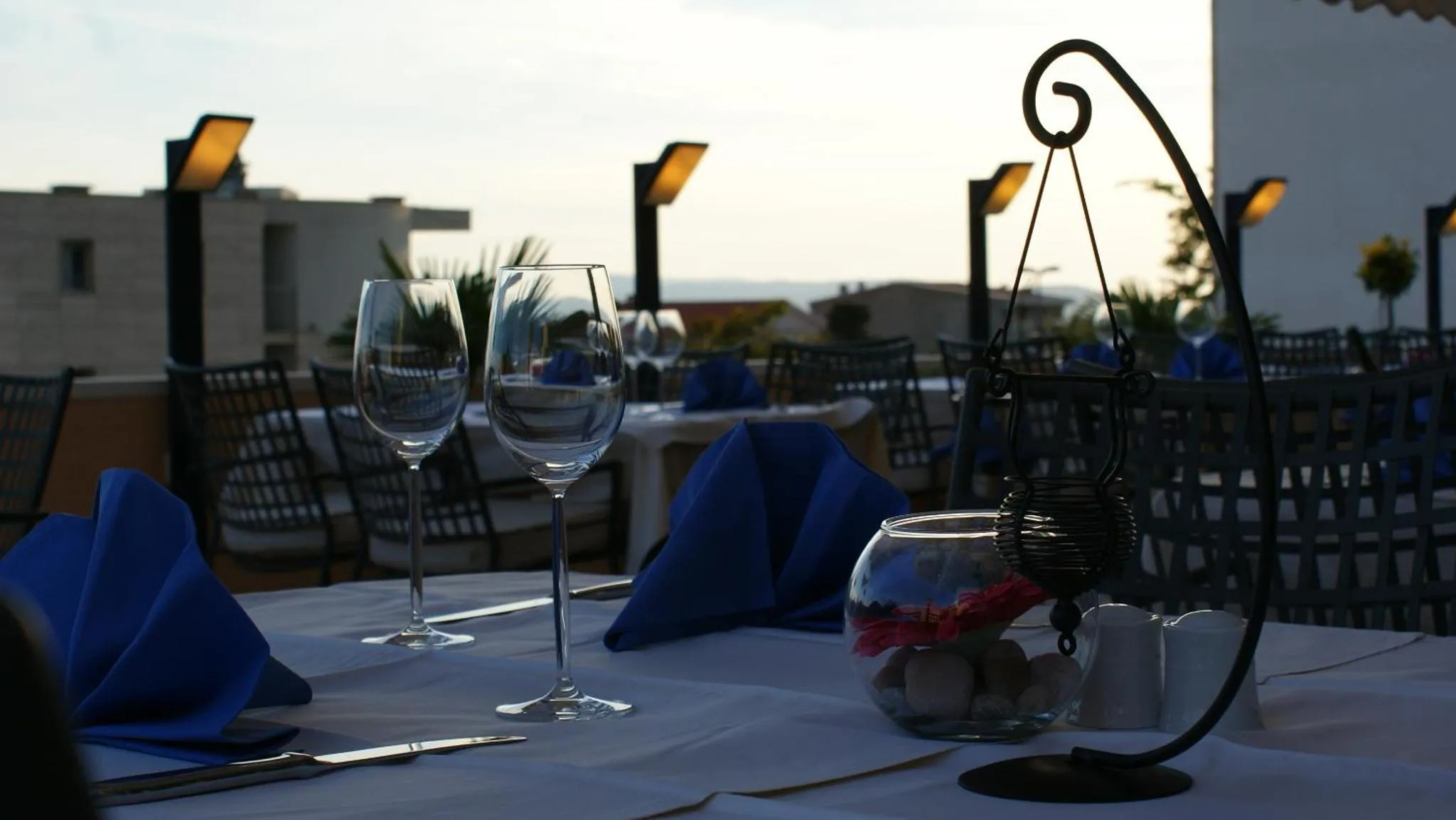 Une table dressée avec des verres à vin et des serviettes bleues sur une terrasse éclairée.