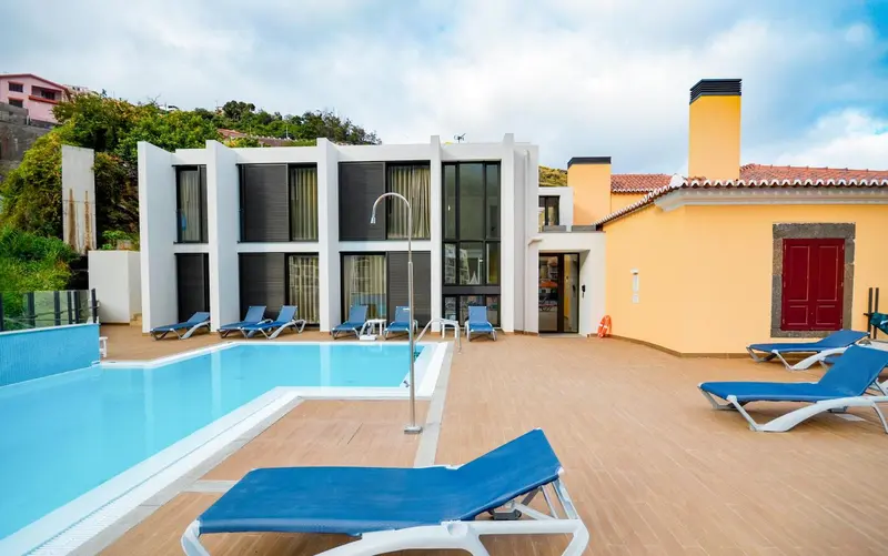 Transats bleus alignés au bord d'une piscine ensoleillée devant un bâtiment blanc.