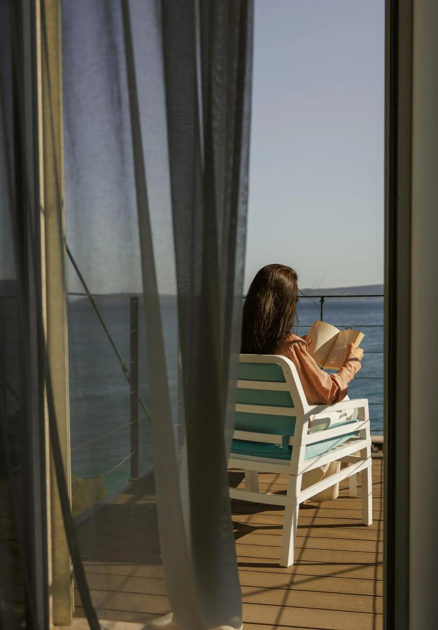 Une femme lit un livre assise sur une terrasse en bois avec vue sur la mer.