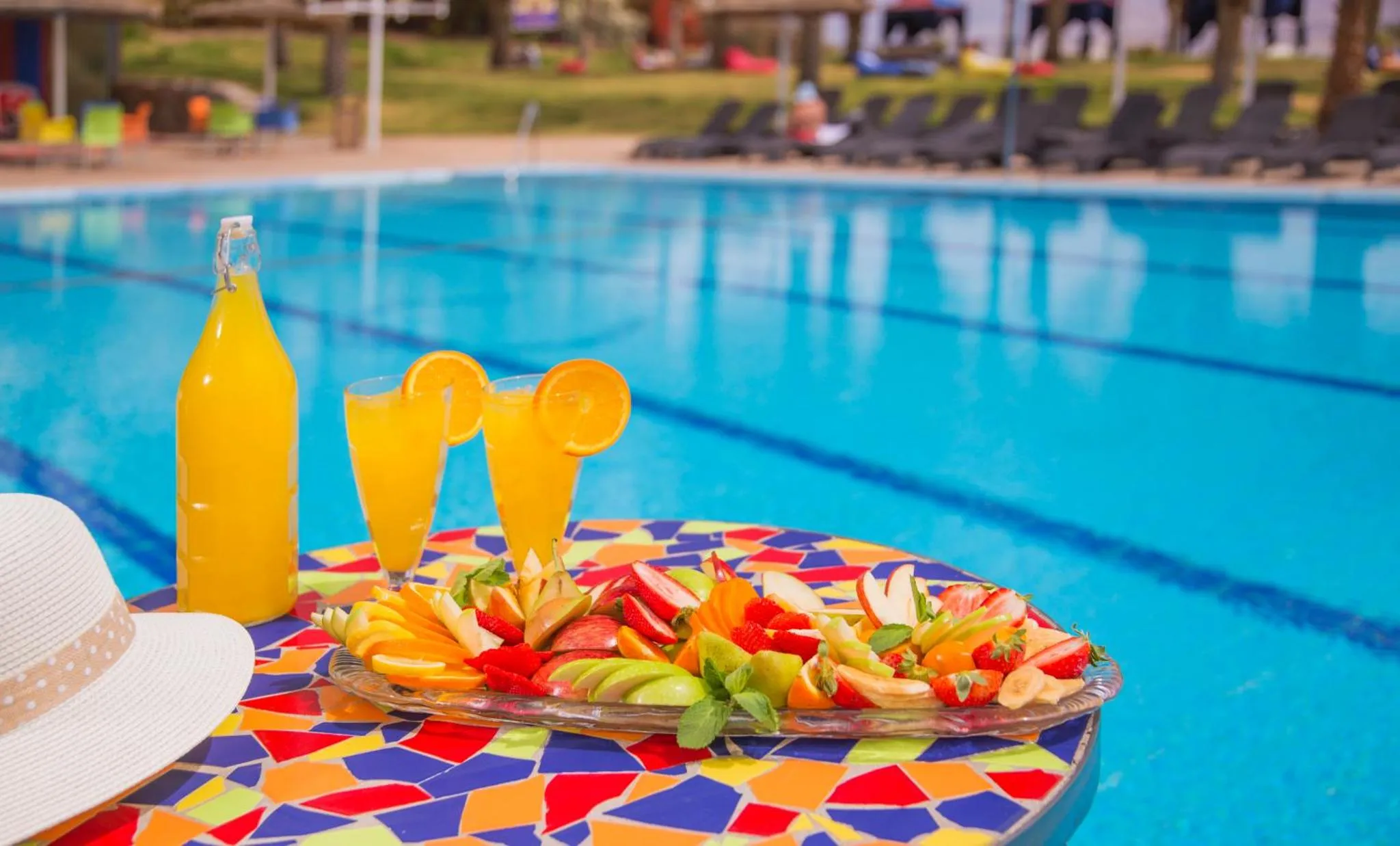 Une table colorée propose des boissons et des fruits frais au bord d'une piscine turquoise.