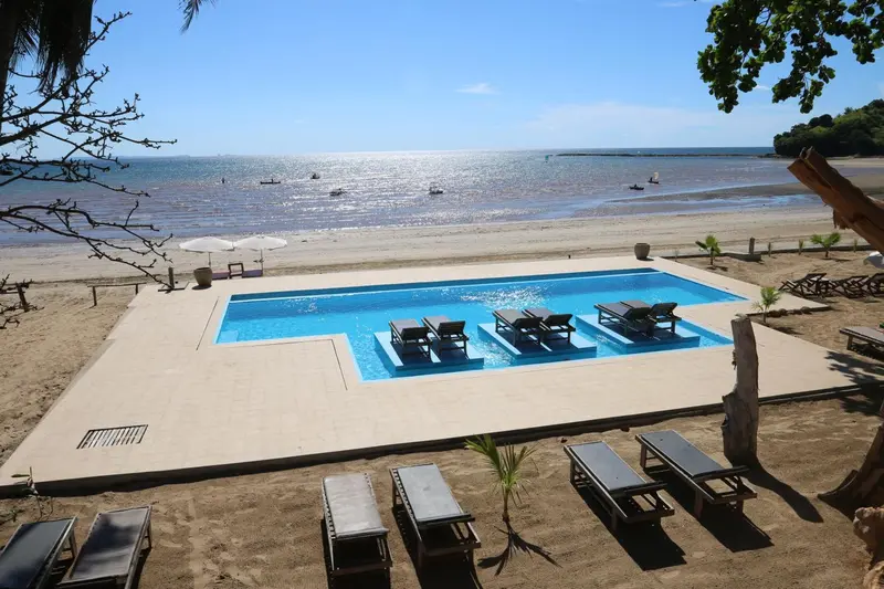 Une piscine rectangulaire bordée de chaises longues se trouve sur une plage de sable fin.