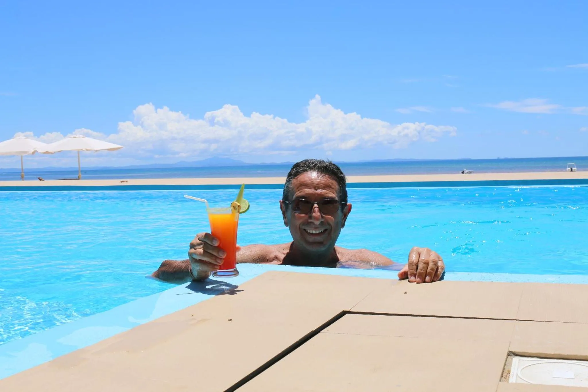 Un homme souriant tient deux cocktails colorés au bord d'une piscine.