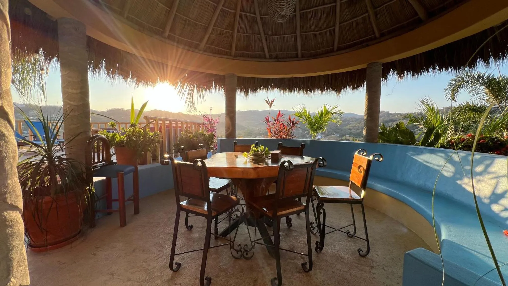 Une table ronde en bois est entourée de sièges sur une terrasse ombragée avec vue sur un paysage verdoyant.
