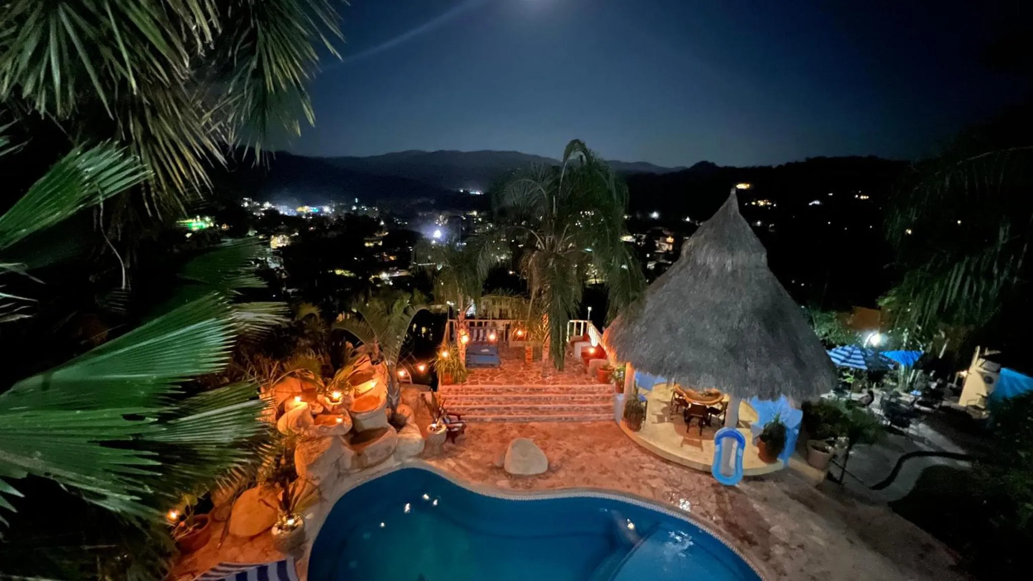 Vue nocturne sur une piscine et un espace de détente avec végétation luxuriante et lumières.