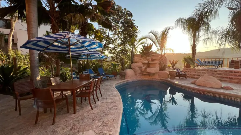 Une piscine bordée d'une terrasse avec tables et chaises, entourée de végétation luxuriante.