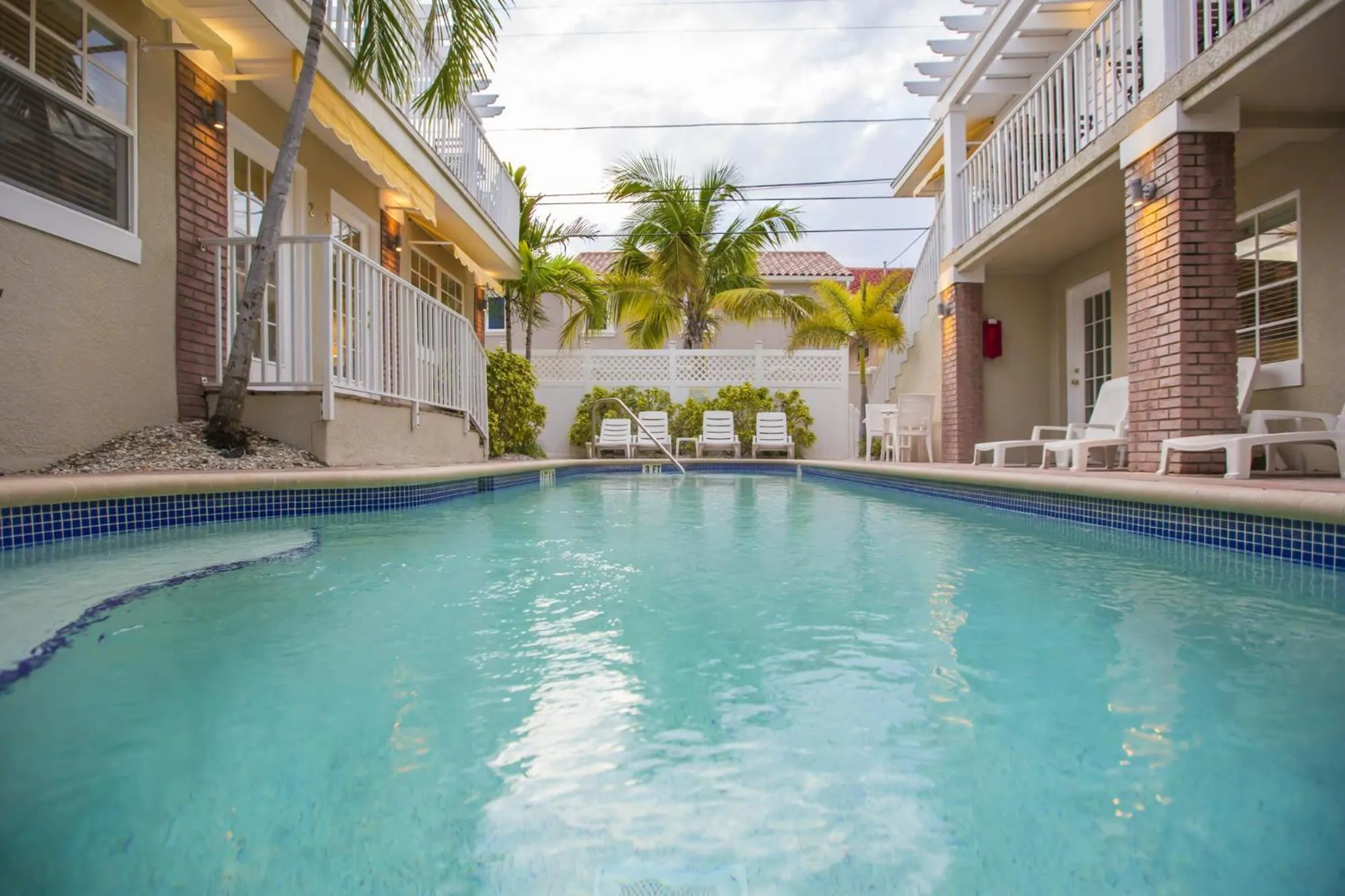 Une piscine rectangulaire turquoise est entourée d'une cour intérieure avec des chaises longues et des palmiers.