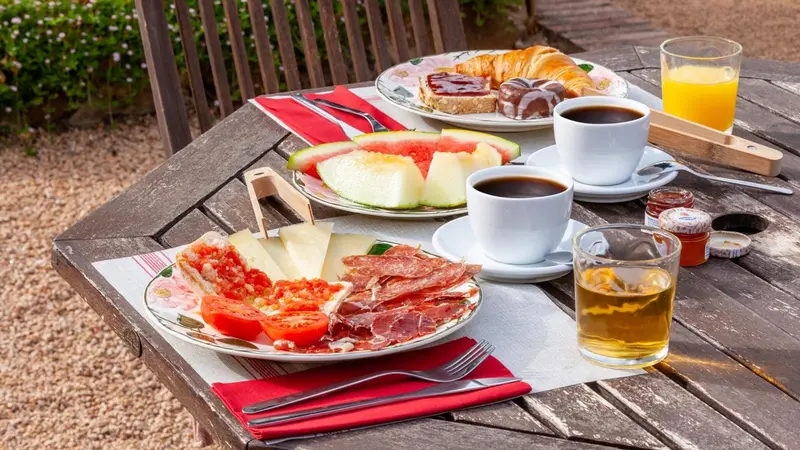 Une table en bois présente un petit-déjeuner avec des fruits, de la charcuterie et des viennoiseries.