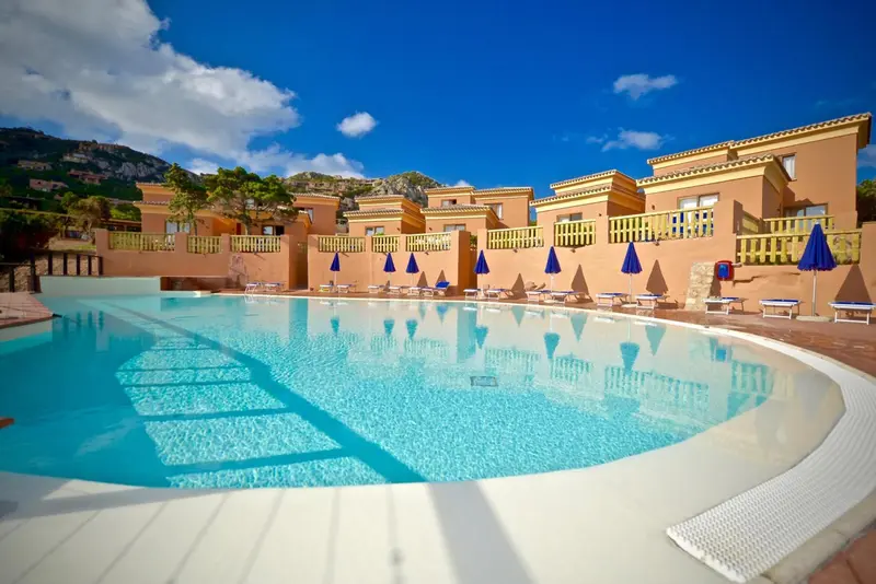 Piscine turquoise bordée de transats devant un bâtiment aux tons pastel.