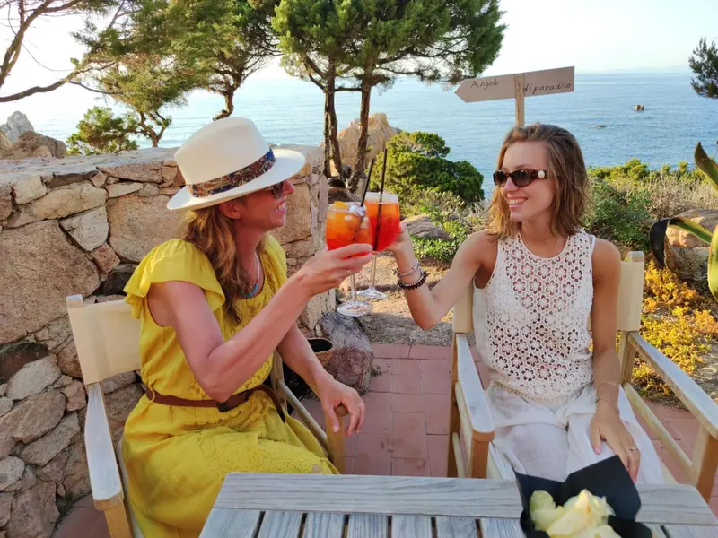Deux femmes sourient en trinquant sur une terrasse ensoleillée avec vue sur la mer.