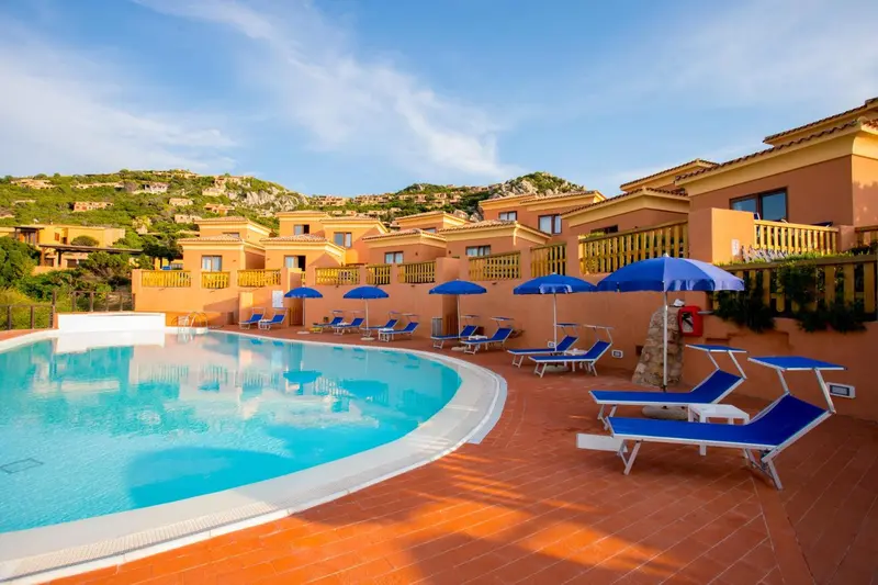 Une piscine turquoise bordée de transats bleus devant un bâtiment aux tons ocre.