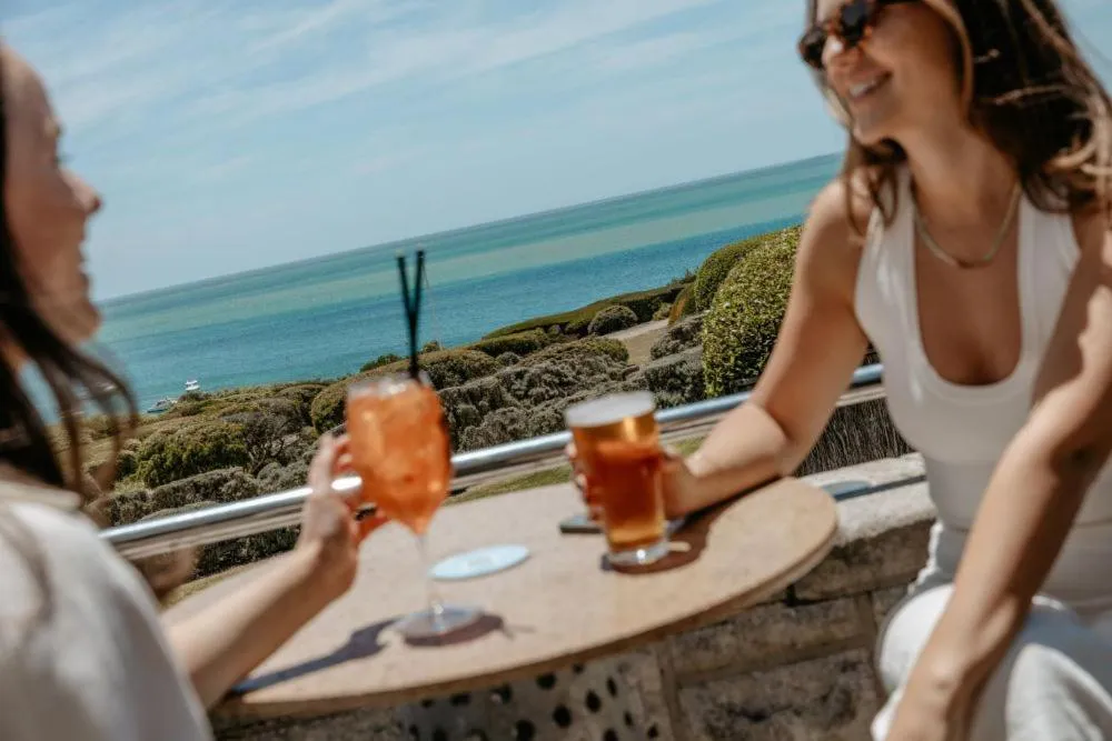 Deux femmes sourient en sirotant des boissons sur une terrasse avec vue sur la mer.